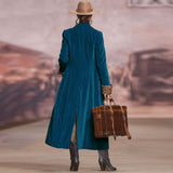 Person in a long blue coat and hat holding a suitcase, walking away with a vintage car and landscape in the background.