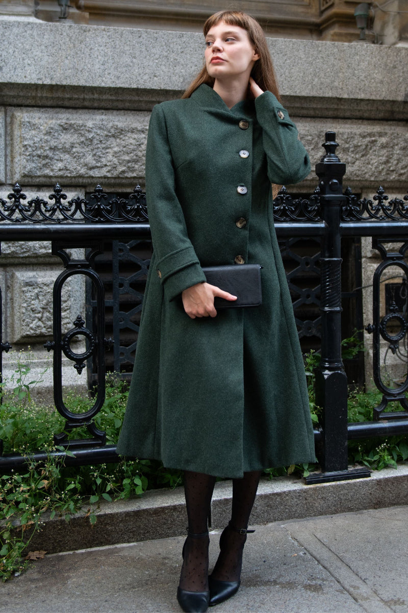 Woman wearing a green coat standing in an urban setting with a black railing and stone wall.