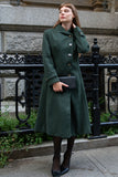 Woman wearing a green coat standing in an urban setting with a black railing and stone wall.