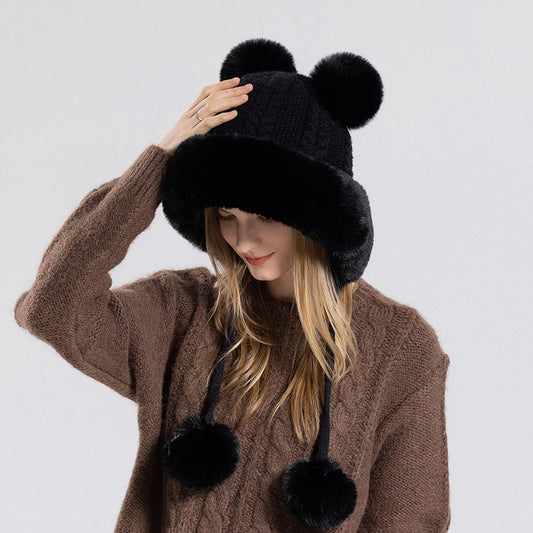 Person wearing a black knitted hat with pom-poms and a brown sweater on a light gray background