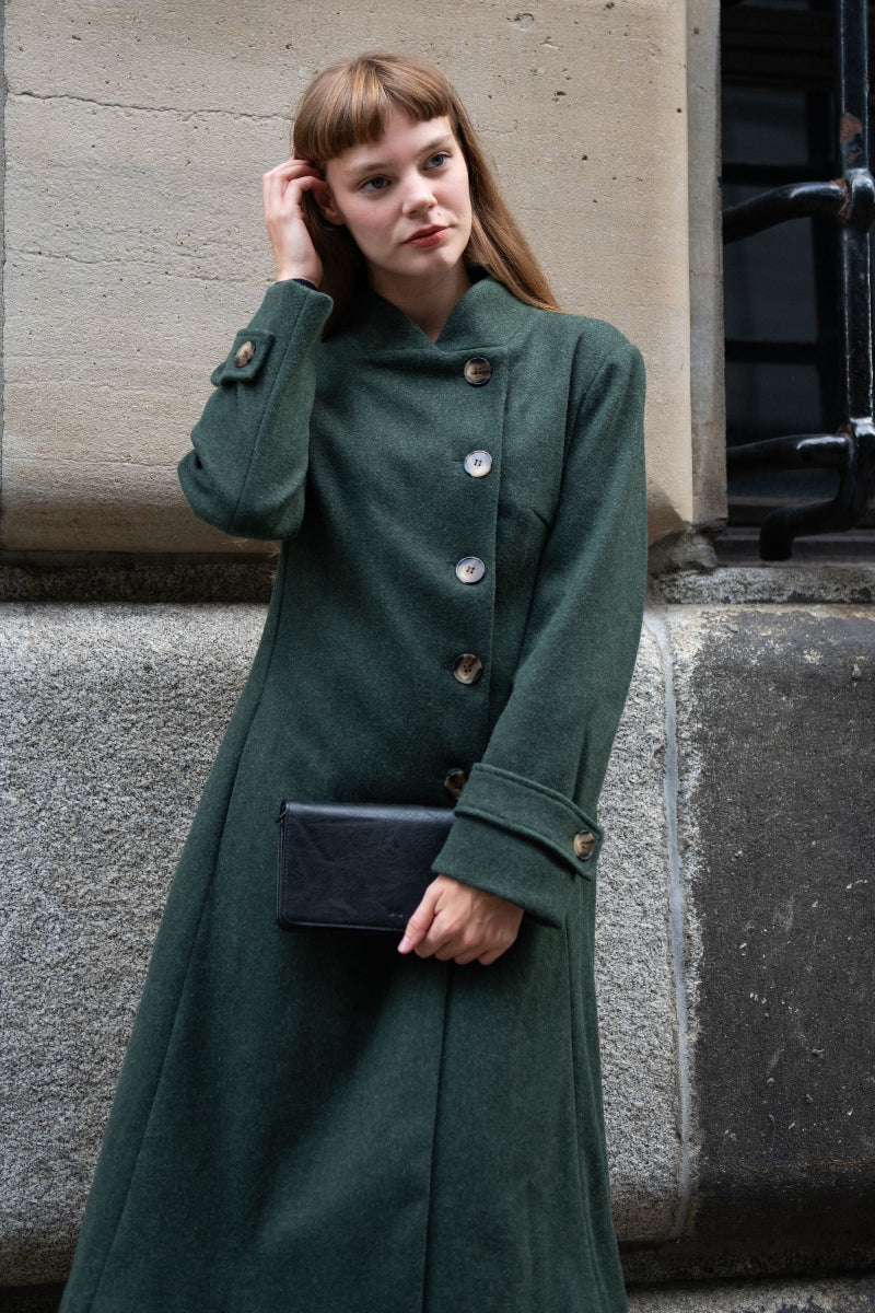 Woman wearing a long green coat holding a black clutch against a neutral background