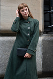 Woman wearing a long green coat holding a black clutch against a neutral background