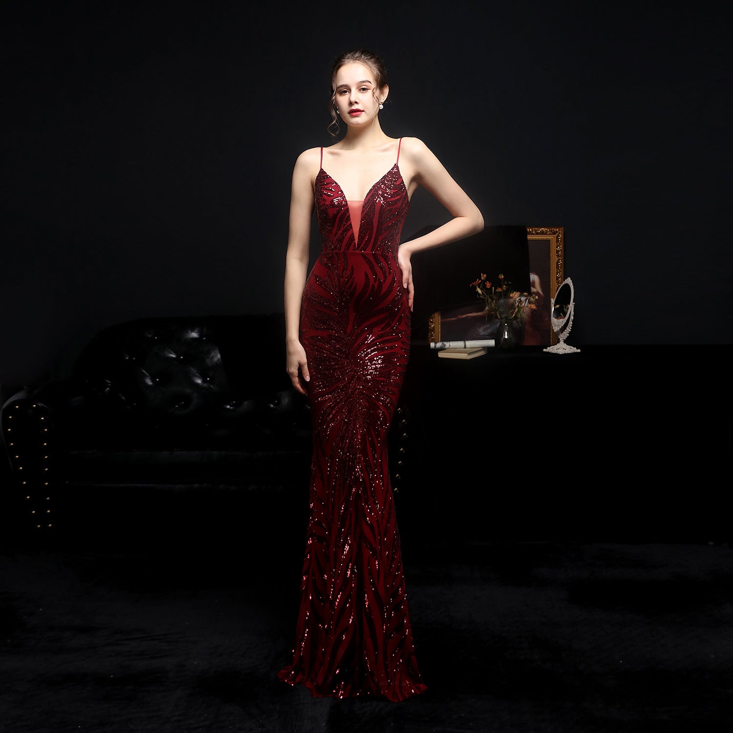 Woman in a red evening gown standing in a dark room with a black sofa and framed picture in the background.