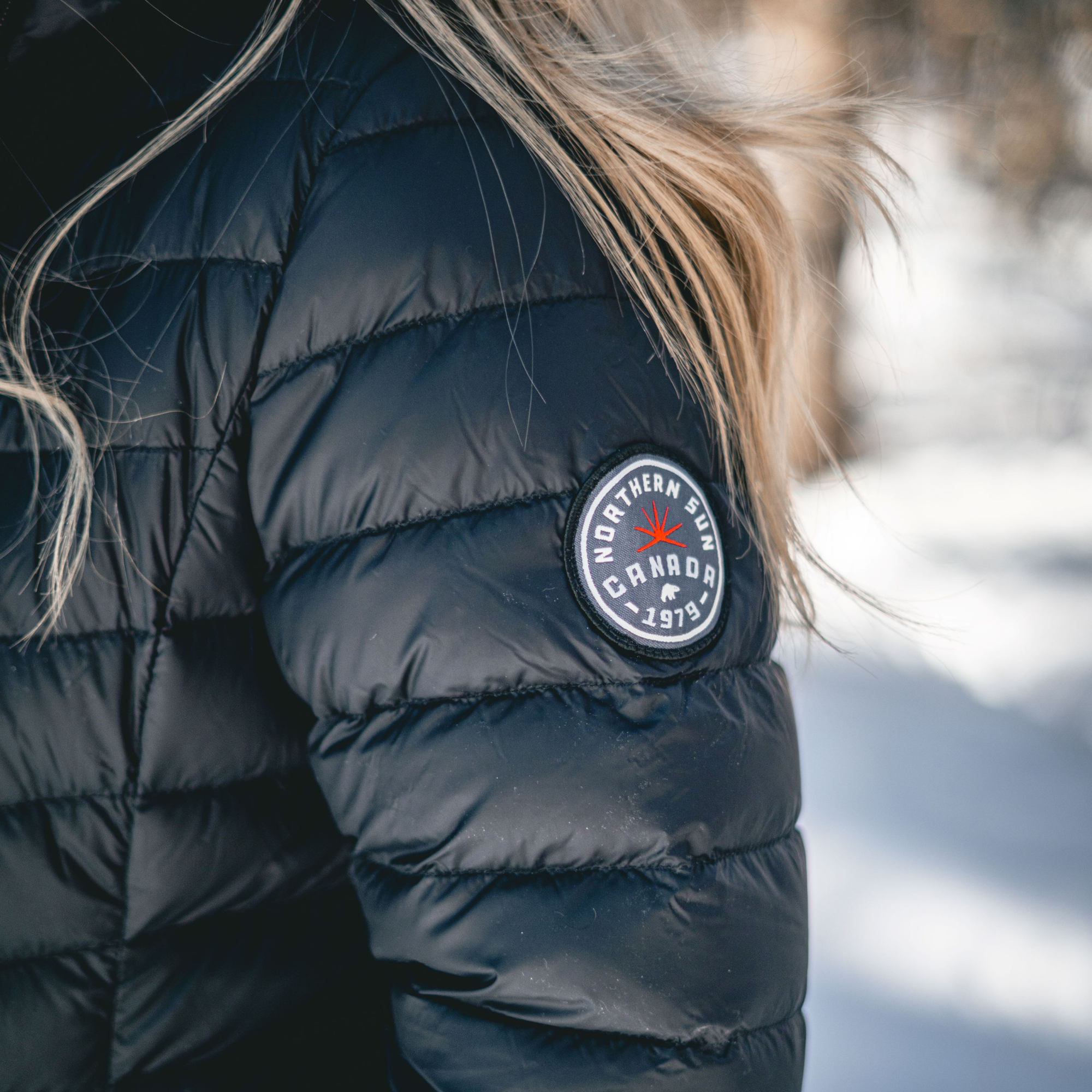 Black puffer jacket with a brand logo on the sleeve against a blurred snowy background